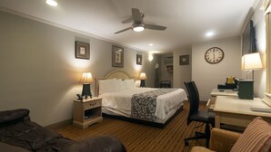 Une chambre d’hôtel comprenant un lit, deux tables de chevet avec des lampes, un bureau avec une chaise, une horloge au mur et un ventilateur de plafond.