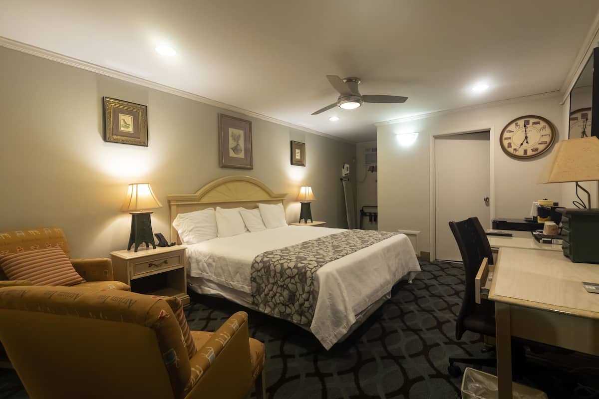 A hotel room with a bed, two bedside tables with lamps, a desk with a chair, a ceiling fan, and a clock on the wall.