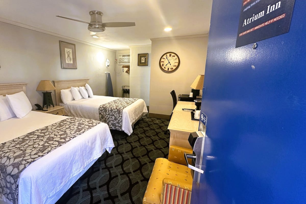 A hotel room with two beds, a desk, a chair, a clock, and a blue wall with an emergency sign.