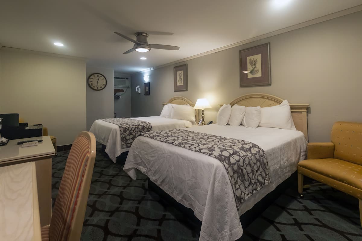 A hotel room with two beds, a desk, a chair, a desk lamp, a clock, and a ceiling fan.