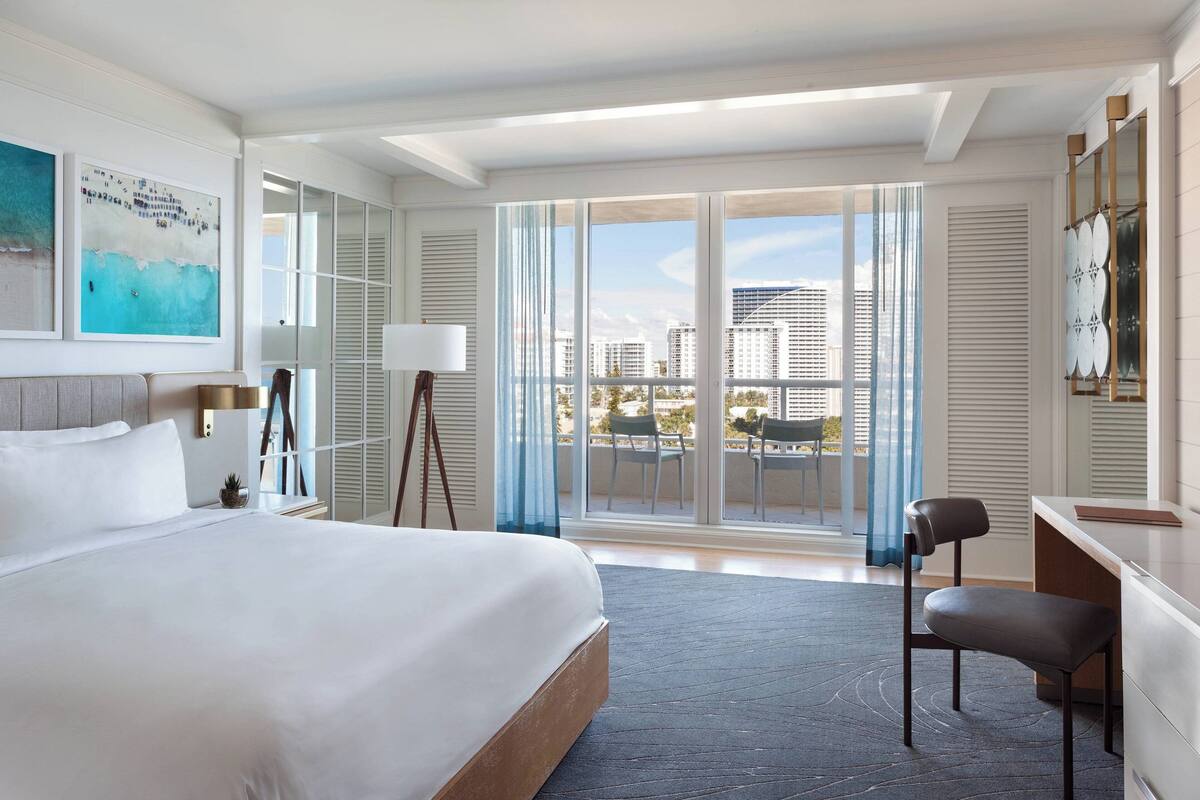A modern hotel room with a large bed, a desk, and a view of the city through floor-to-ceiling windows.