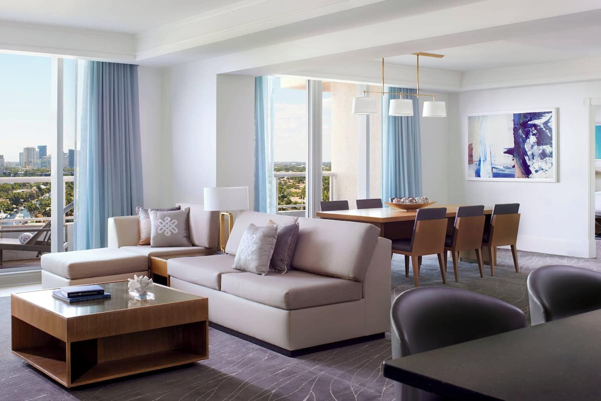 A modern hotel room with a sofa, dining table, chairs, and a city view.