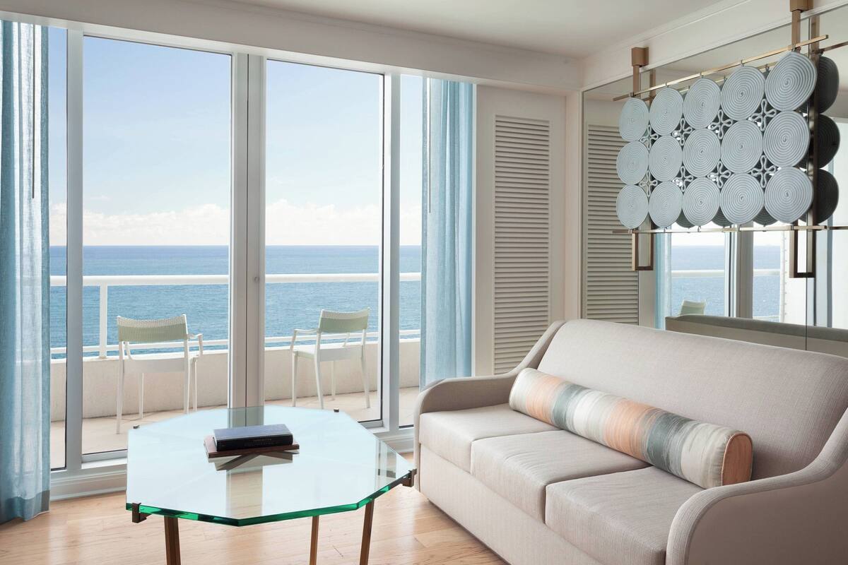 A modern living room with a glass table, a beige sofa, and a view of the ocean through large windows.