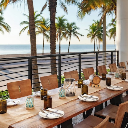 A long wooden dining table set for a meal with chairs, placed on a balcony overlooking a beach with palm trees and the ocean.