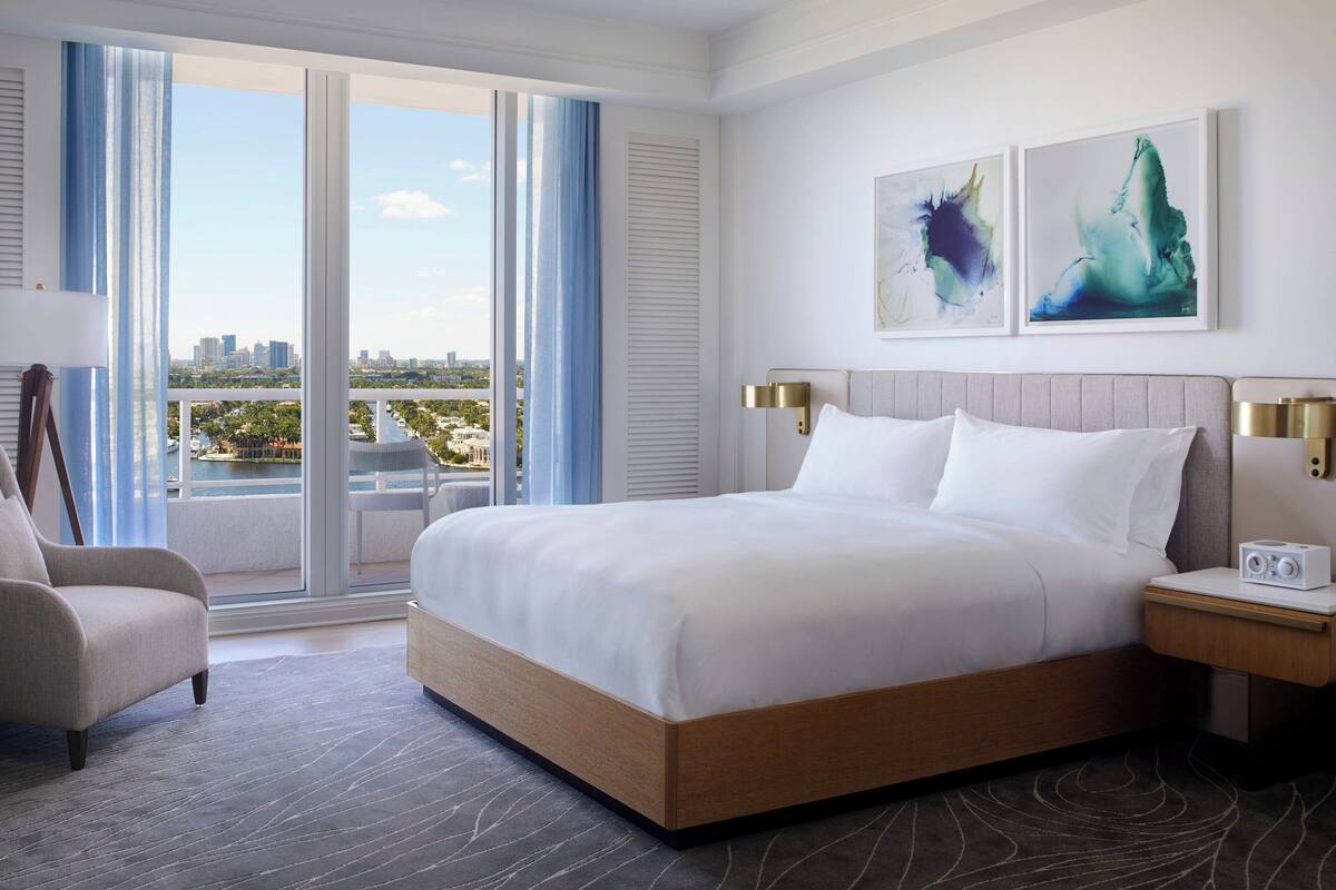 A hotel room with a large bed, a chair, a nightstand, and a view of the city through large windows.