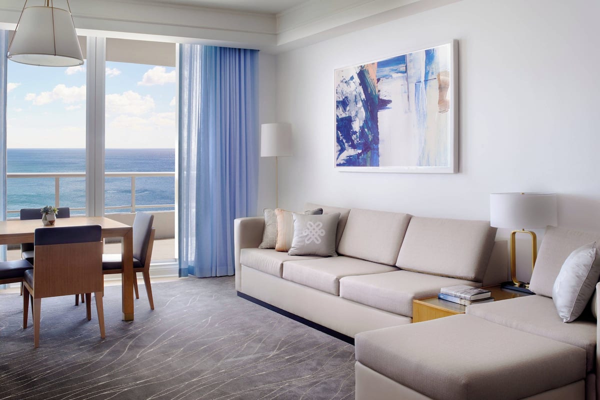 A modern hotel room with a large window offering a view of the ocean, a sofa, a dining table with chairs, and a painting on the wall.