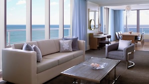 A modern hotel room with a large balcony, a sofa, a dining area, and a view of the ocean.