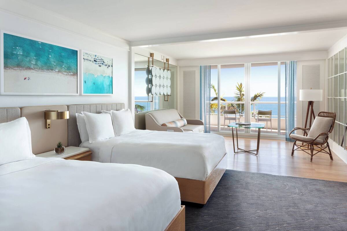 A hotel room with two beds, a balcony with a view of the sea, and a seating area with a table and chairs.