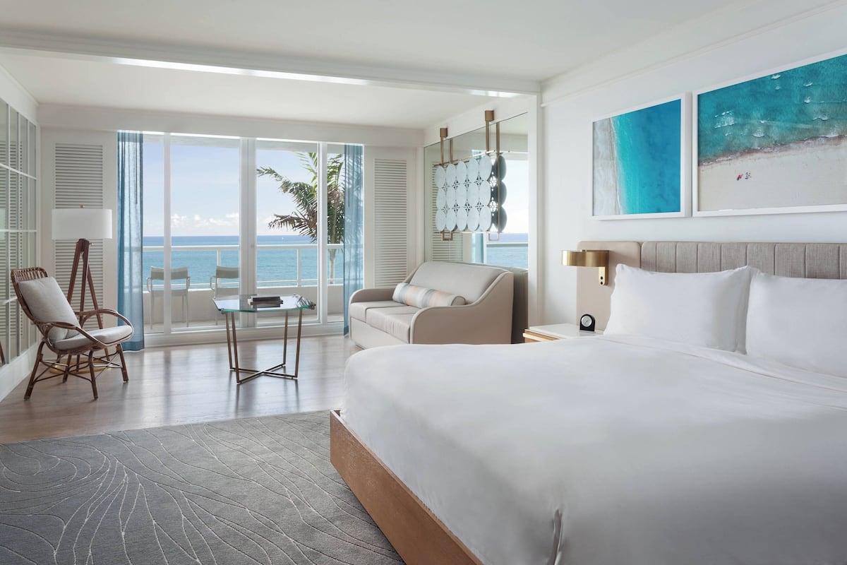 A hotel room with a large bed, a sofa, a chair, a small table, and a view of the ocean through large windows.