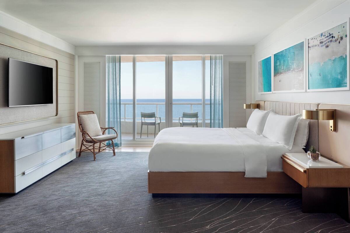 A modern hotel room with a large bed, a flat-screen TV, a balcony with a view of the sea, and a dining area with chairs.