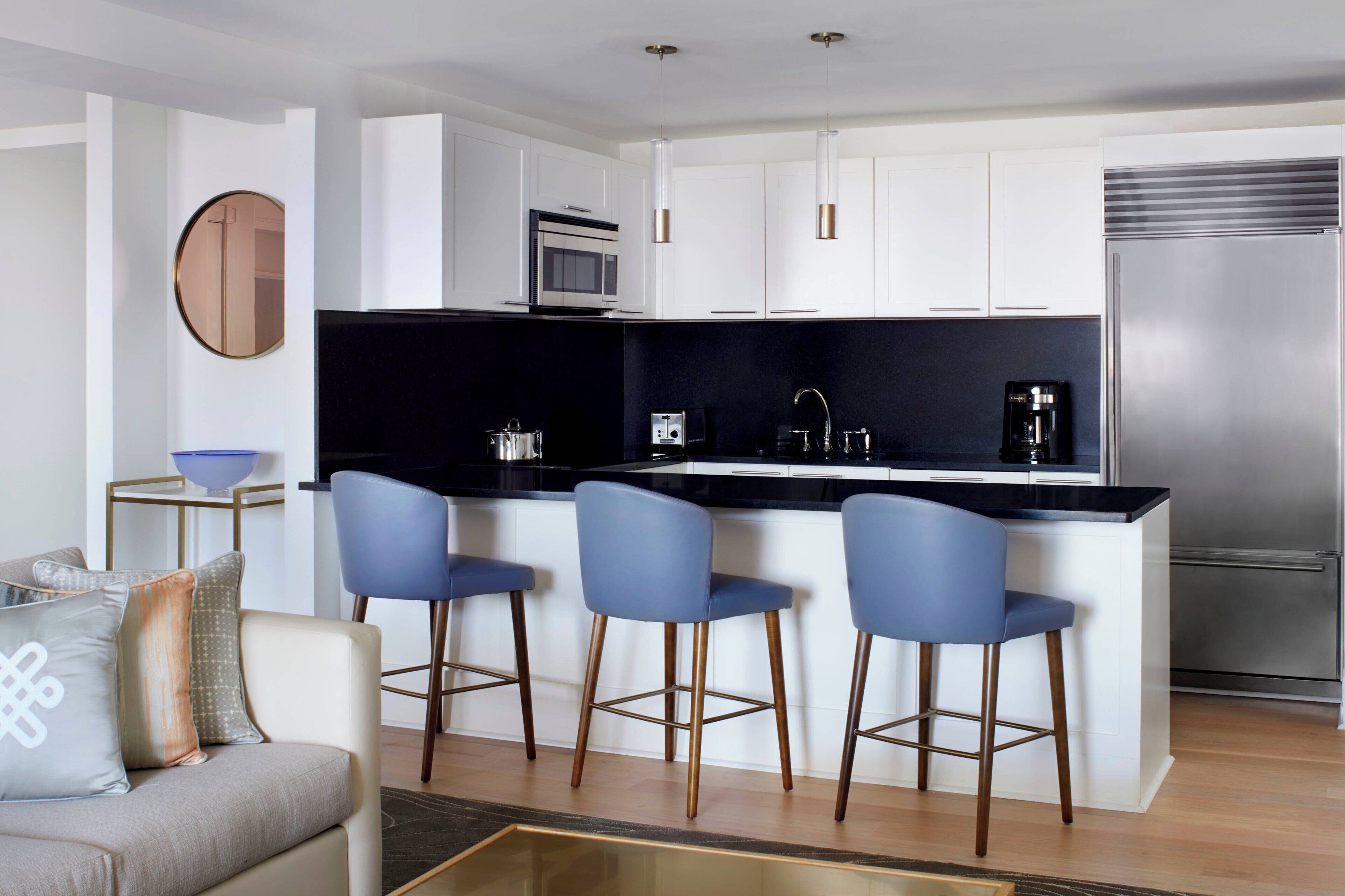 Een moderne keuken met witte kasten, een zwart aanrecht en een roestvrijstalen koelkast. Drie blauwe barkrukken en een koffieapparaat zijn aanwezig.