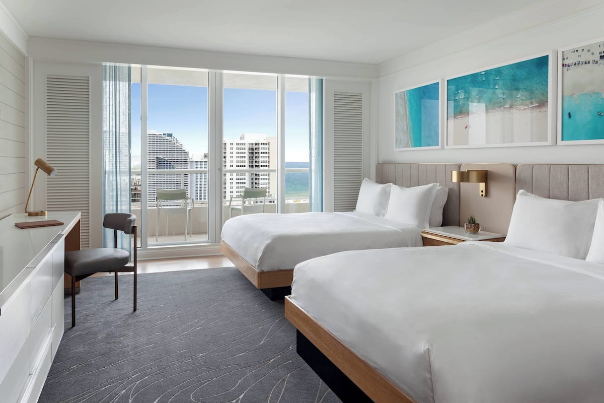 A hotel room with two beds, a desk, a chair, and a view of the city.
