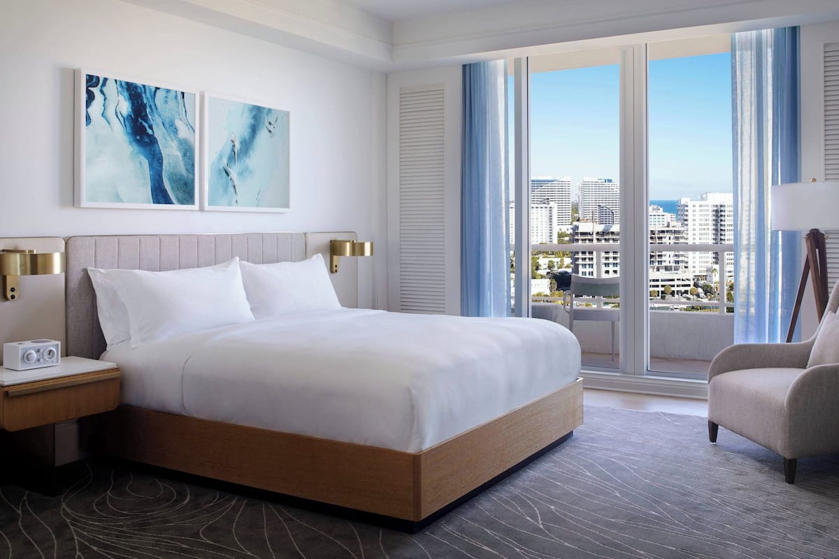 A hotel room with a large bed, a nightstand with a lamp, a chair, and a view of the city through large windows.