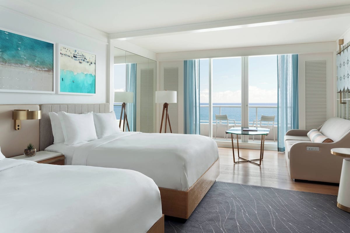 A hotel room with two beds, a seating area with a table, and a large window offering a view of the sea.