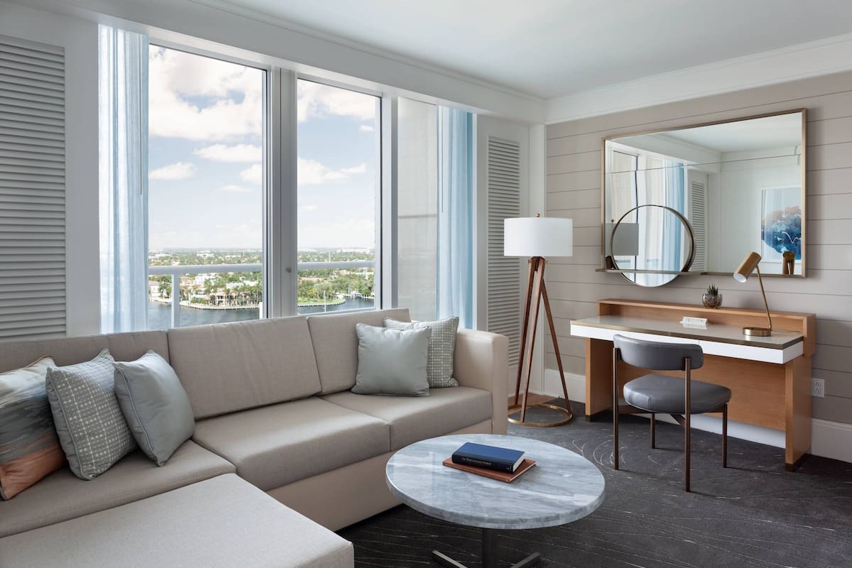 A modern hotel room with a sofa, a desk, a round table, and a large window offering a city view.