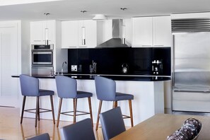 A modern kitchen with white cabinets, a black countertop, and stainless steel appliances. There are three bar stools and a wooden dining table.