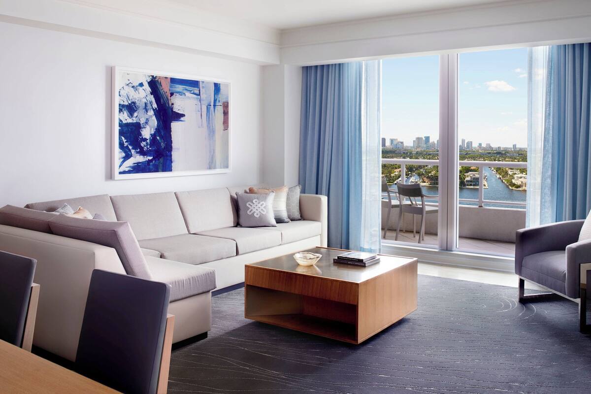 A modern living room with a sofa, a coffee table, and a view of a cityscape through large windows.