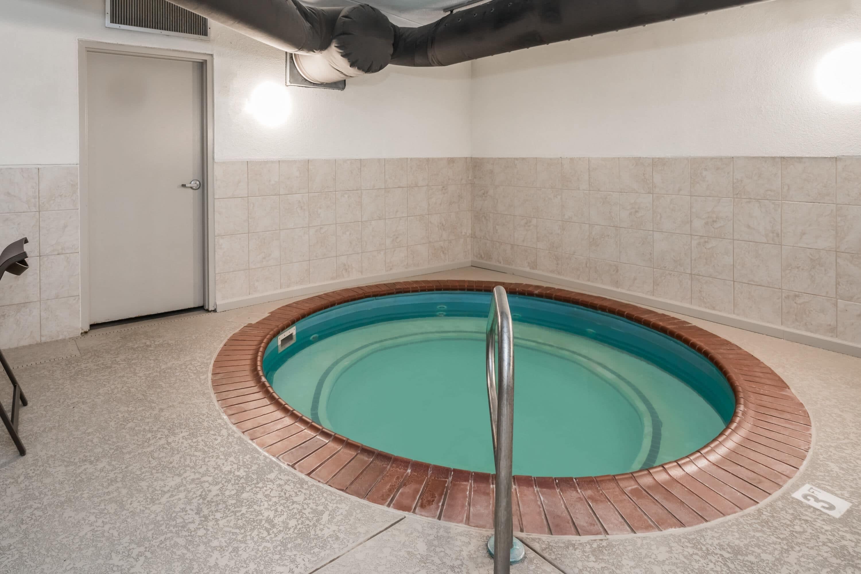 A circular hot tub with a metal railing, surrounded by beige tiles, in a room with a door and a vent on the ceiling.