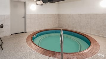 Uma banheira de hidromassagem circular com guarda-corrimão metálico, rodeada por azulejos bege, em um quarto com porta e um respiradouro no teto.