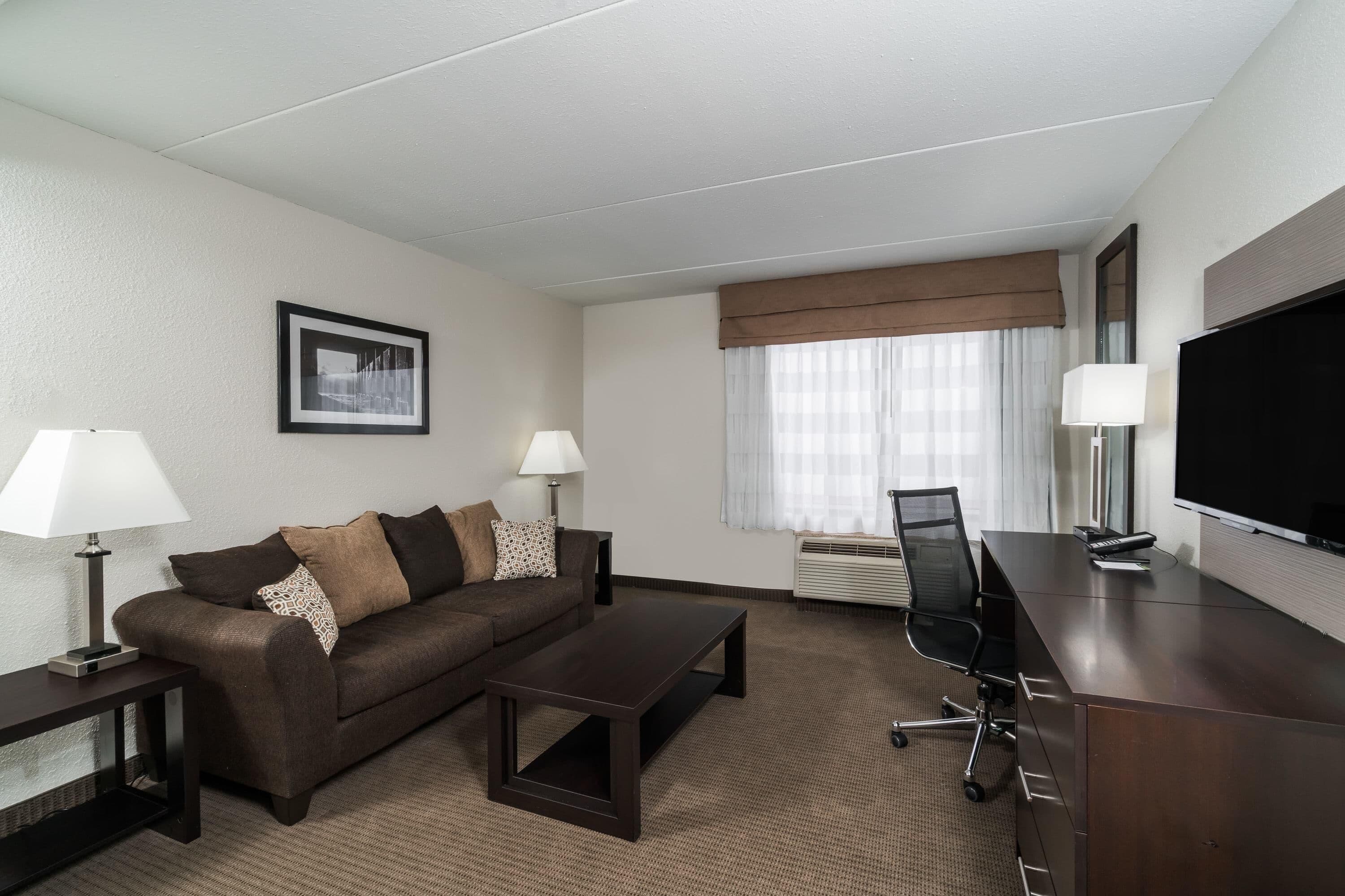 A hotel room with a sofa, a coffee table, a desk with a chair, a TV, and a window with curtains.