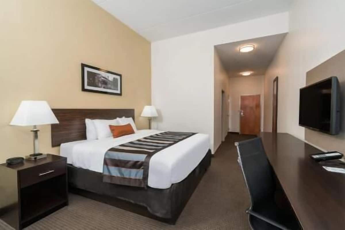 Ein Hotelzimmer mit Bett, Nachttischen, einem Schreibtisch, einem Sessel und einem Fernseher, sowie einem gerahmten Bild an der Wand.