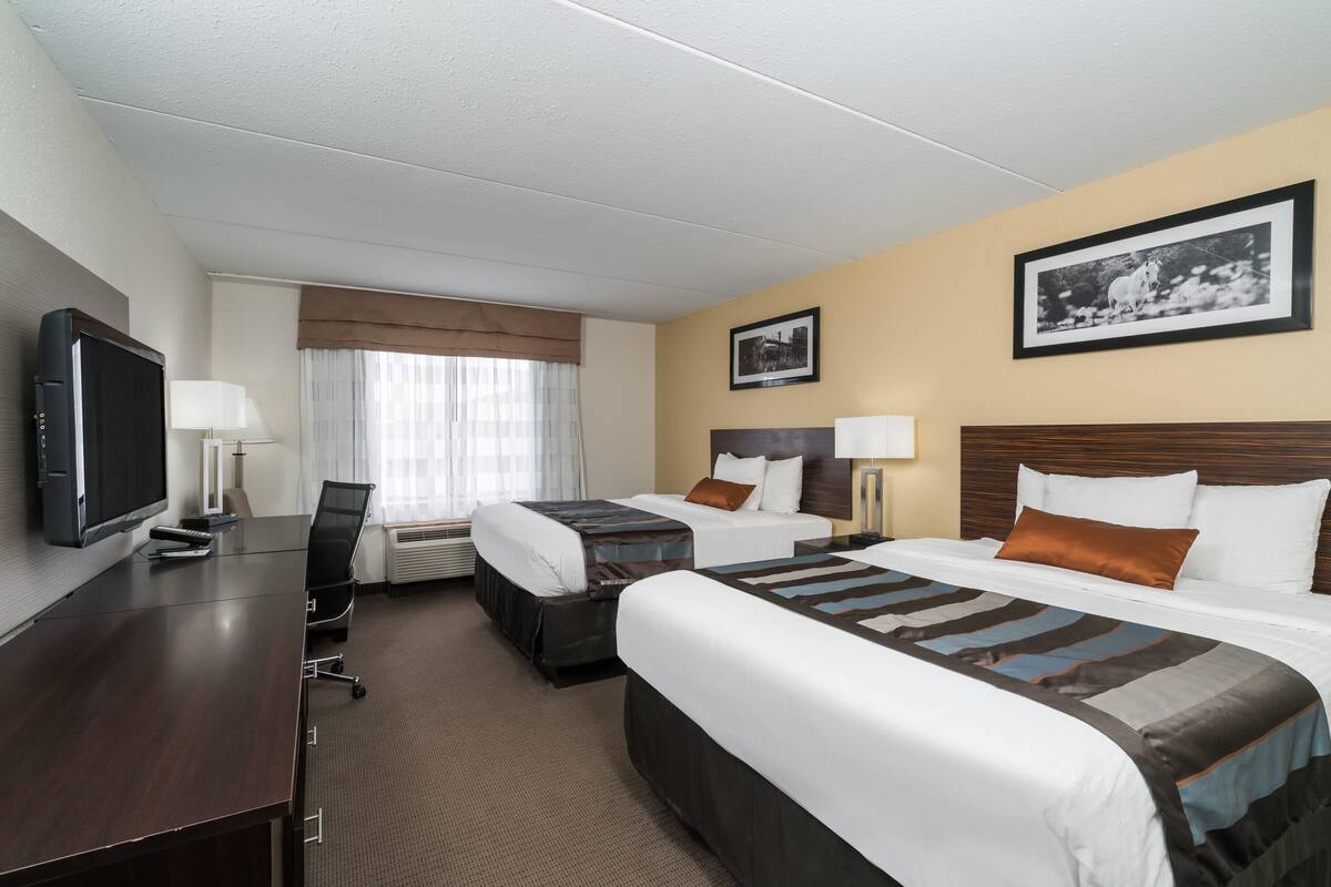 Ein Hotelzimmer mit zwei Betten, einem Schreibtisch mit Fernseher, einem Sessel und einem Fenster mit Vorhängen.