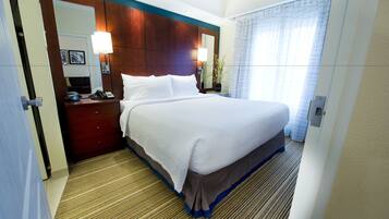 A hotel room with a large bed, a wooden headboard, a nightstand with a lamp, a mirror, and a window with curtains.