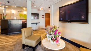 A hotel lobby with a reception desk, a seating area with a chair, a round table with a floral arrangement, and a large flat-screen TV mounted on the wall. - Best Western Nittany Inn Milroy (Reedsville)