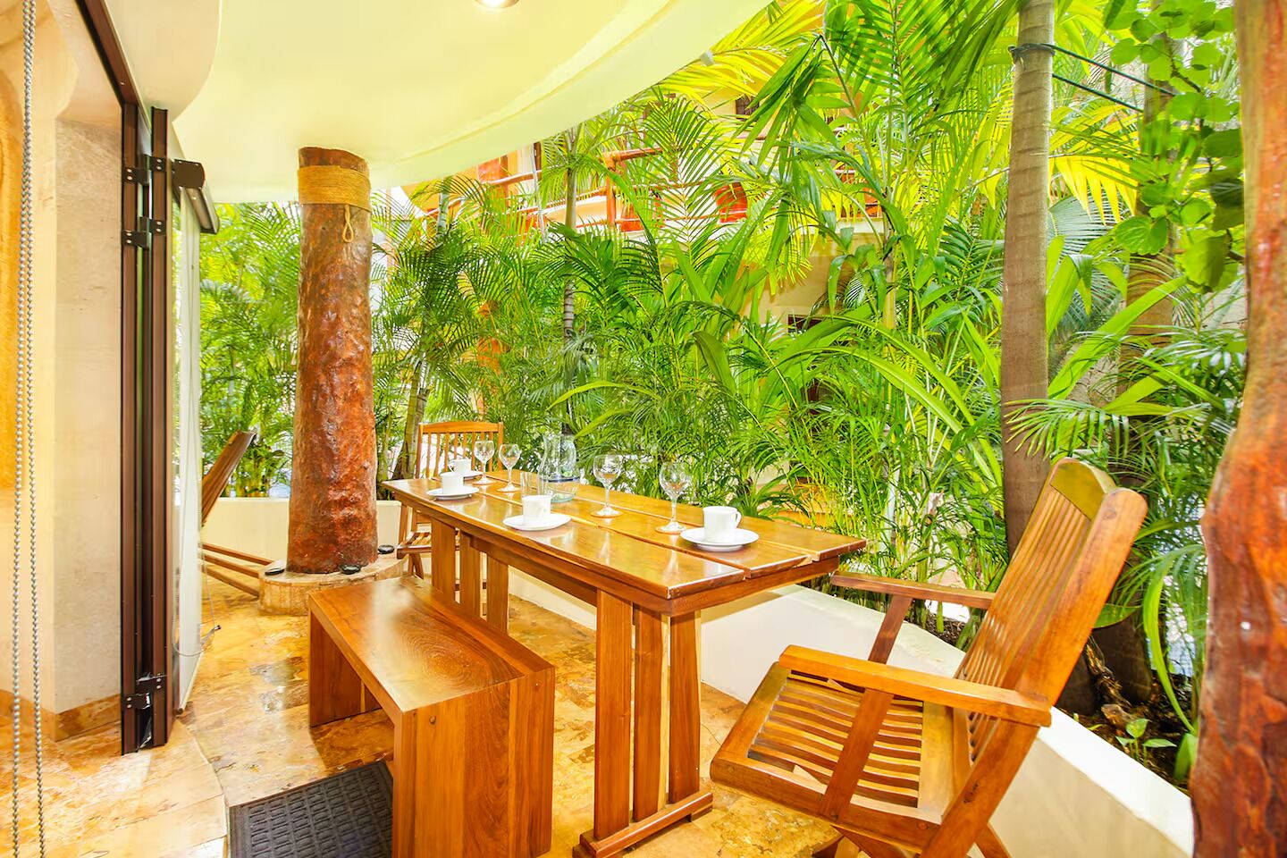 Una mesa de comedor de madera dispuesta con platos y vasos blancos, rodeada de sillas, en una habitación con vistas a un jardín tropical.