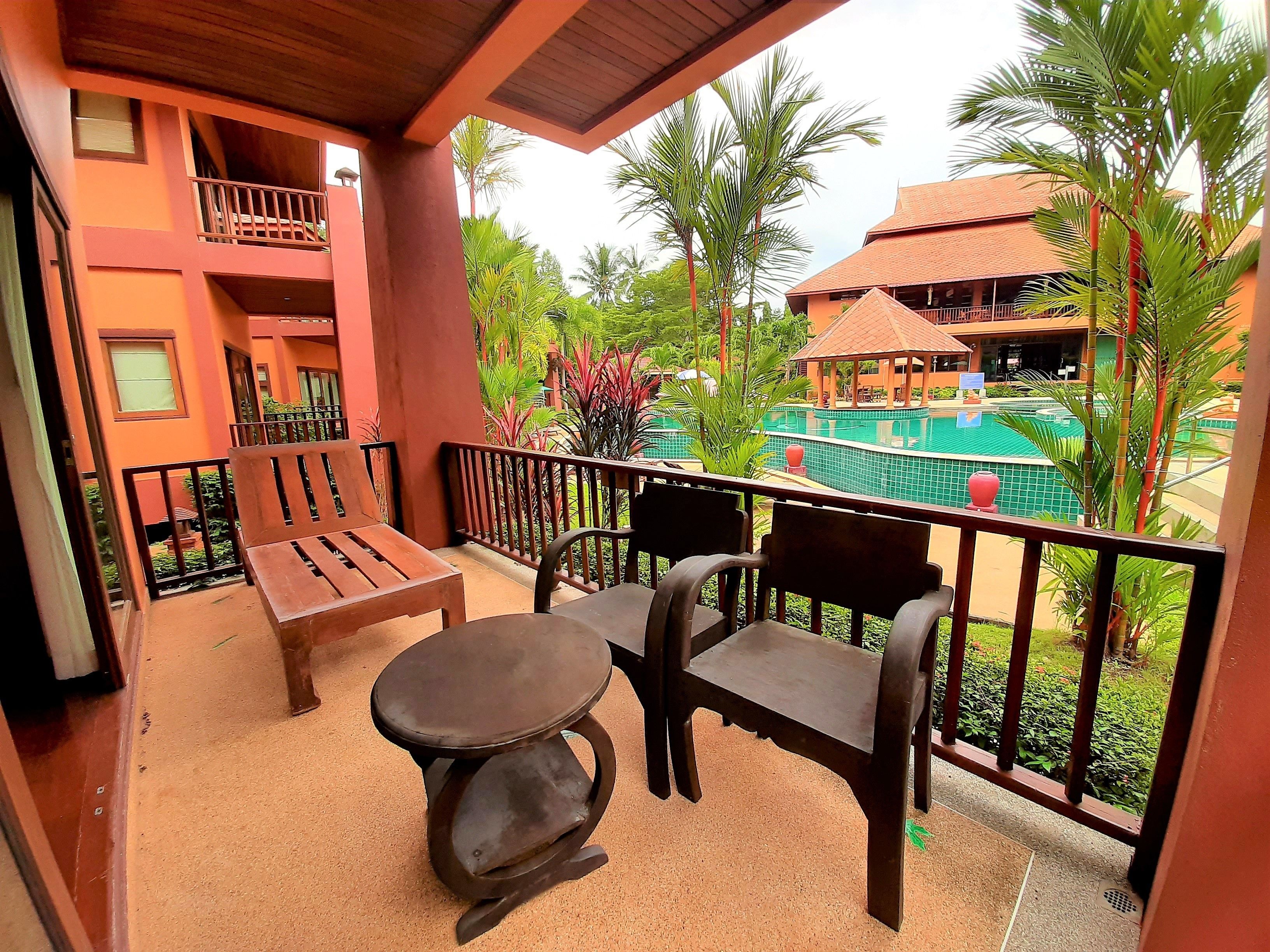 A balcony with wooden furniture, a view of a swimming pool, and surrounding greenery.