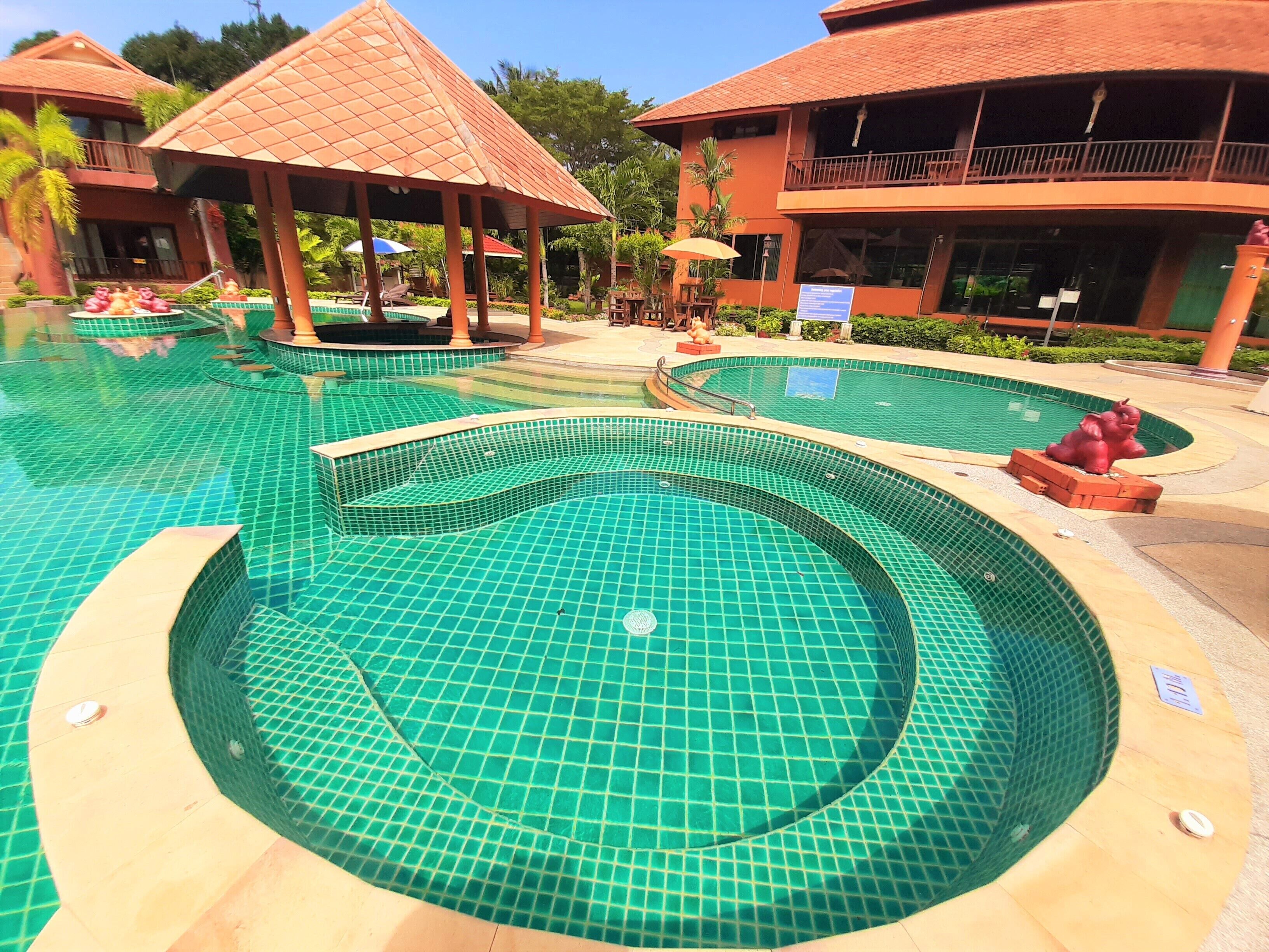 Une piscine dont le fond est recouvert de carreaux, entourée de bâtiments dotés de balcons et d’un pavillon abrité.