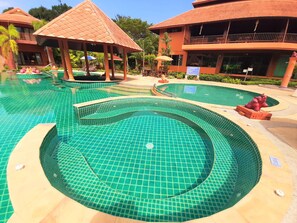 Une piscine dont le fond est recouvert de carreaux, entourée de bâtiments dotés de balcons et d’un pavillon abrité.
