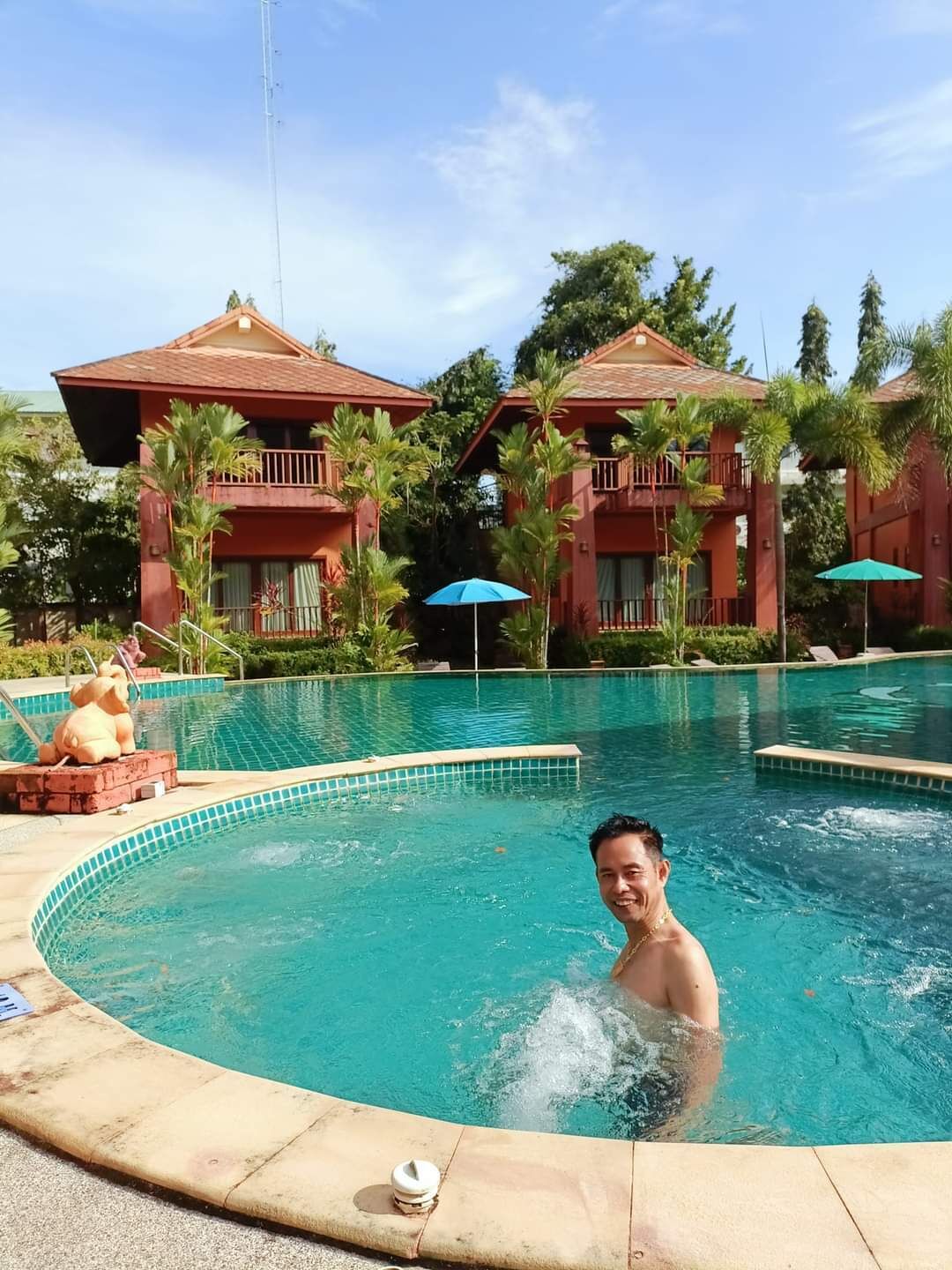 A man in a swimming pool with a poolside lounge area, palm trees, and two red-roofed buildings with balconies.