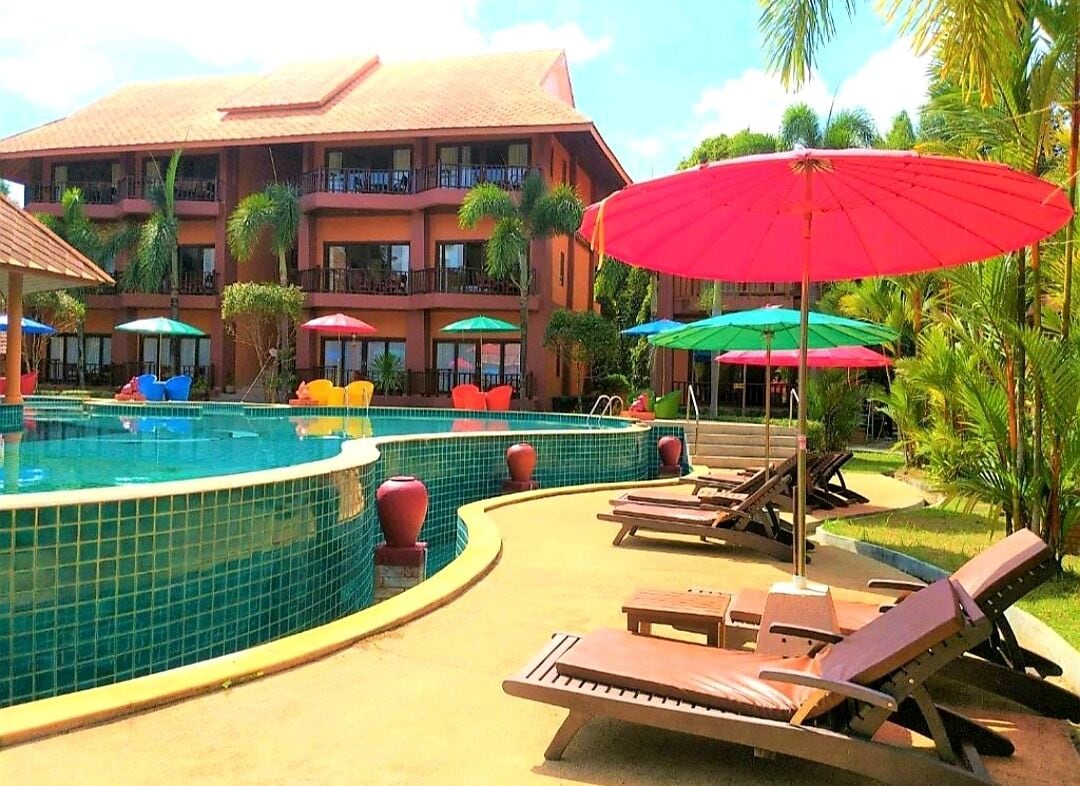 A pool area with lounge chairs and umbrellas, surrounded by a building with balconies and palm trees.