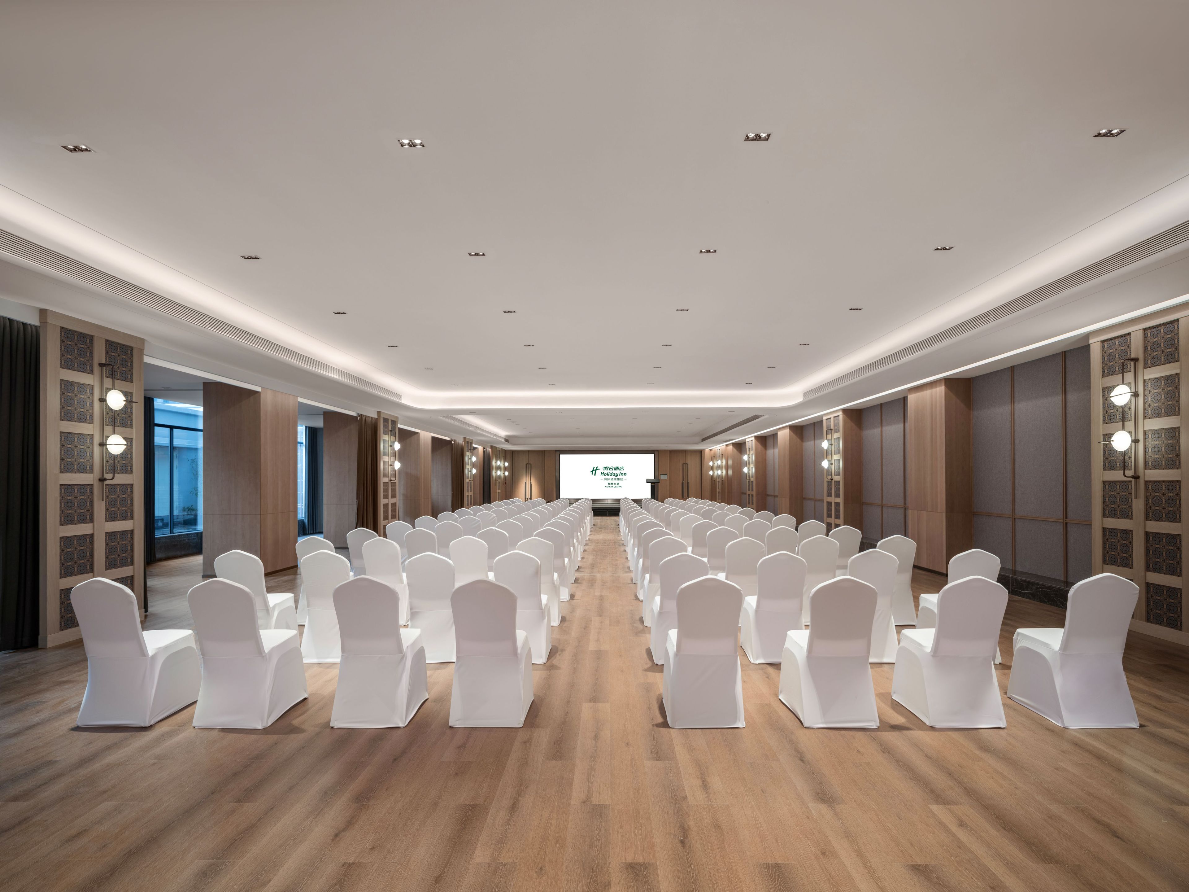 Un salón de conferencias con filas de sillas blancas, una pantalla grande al frente y paneles de madera en las paredes.