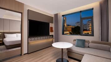 A modern hotel room with a bed, a TV, a sofa, and a view of the cityscape at dusk.