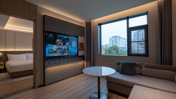 Ein Hotelzimmer mit Bett, Wandfernseher, rundem Tisch, einer Couch und einem großen Fenster mit Blick auf die Stadt.