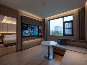 Une chambre d’hôtel avec un lit, un téléviseur fixé au mur, une table ronde, un canapé et une grande fenêtre donnant sur la ville.