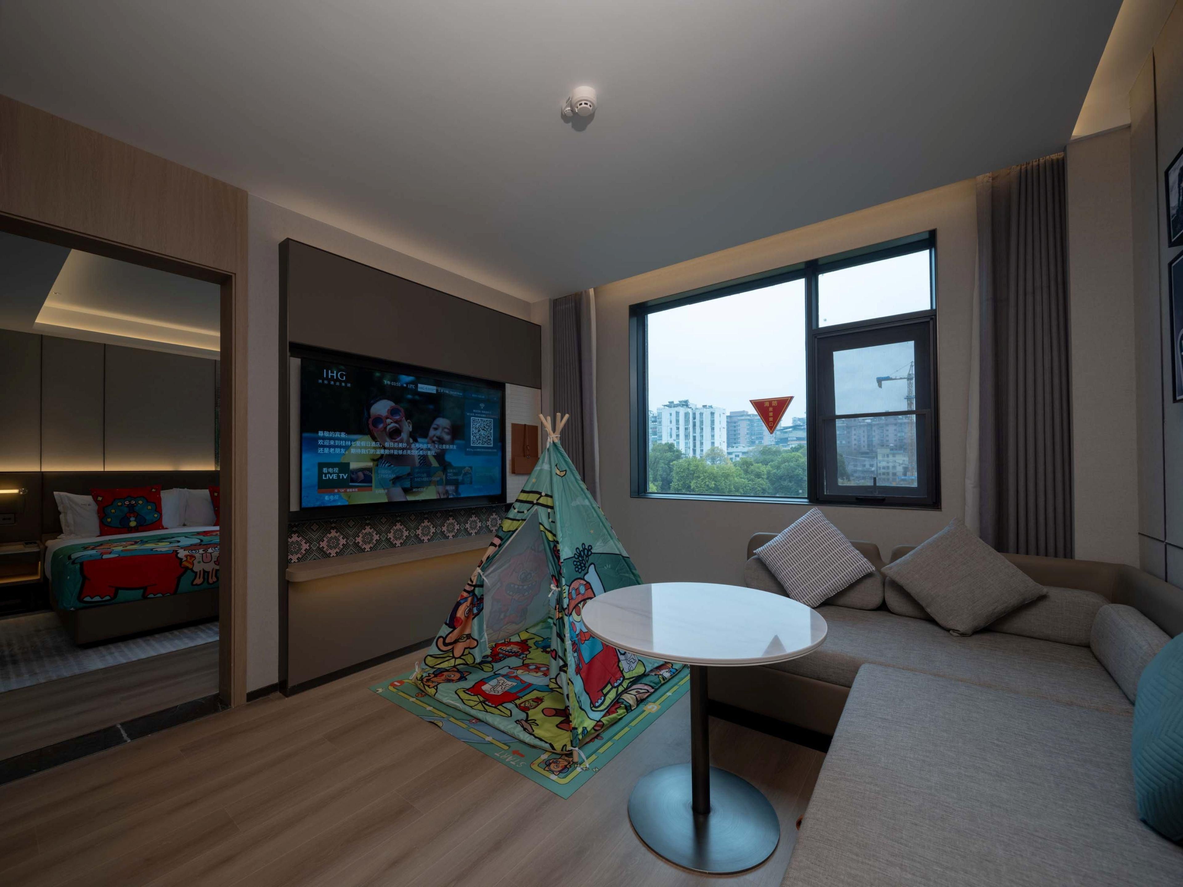Habitación de hotel moderna con un televisor grande, un sofá acogedor, una pequeña mesa redonda y una colorida tienda de juegos para niños.