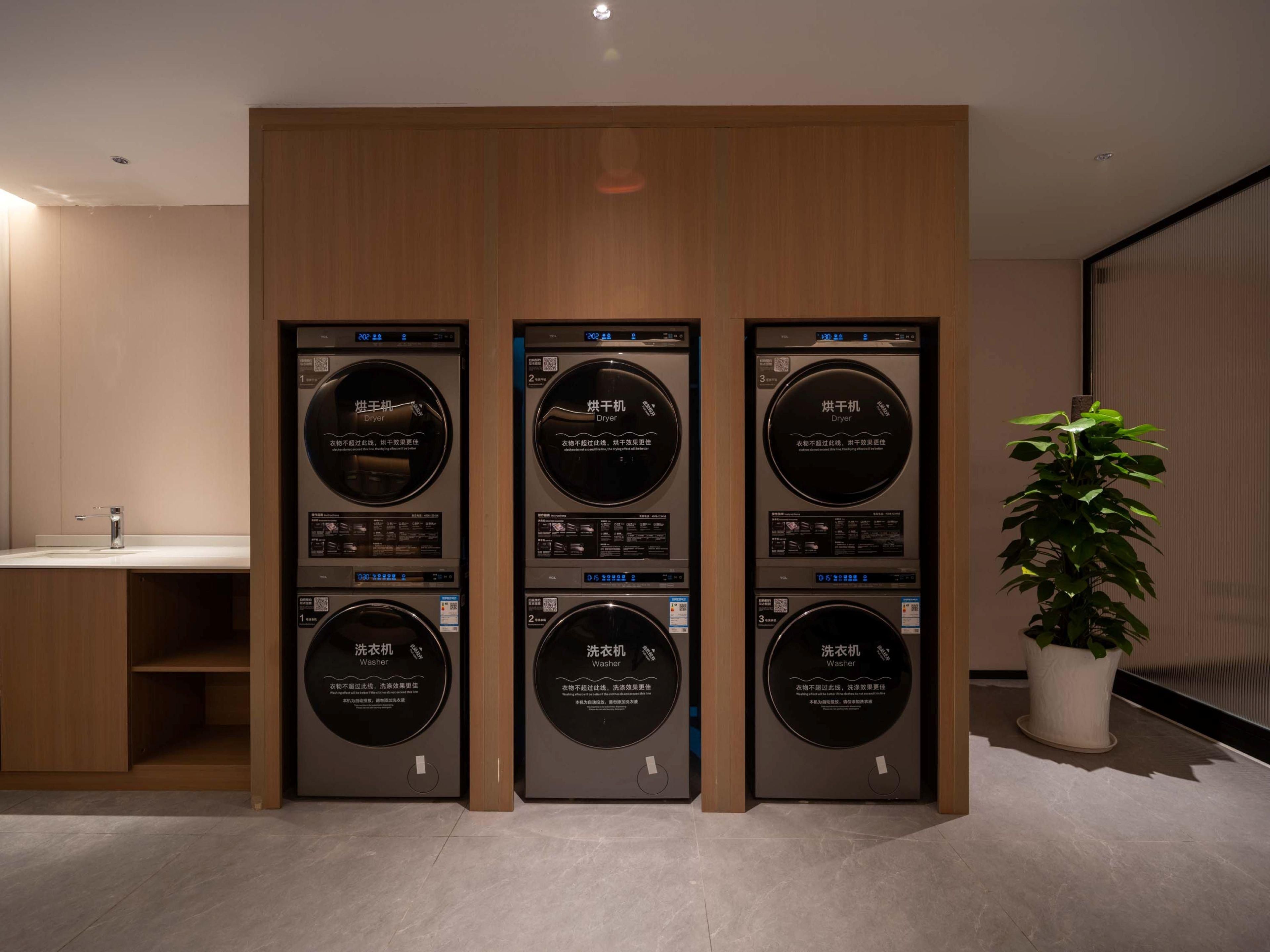 A row of three wall-mounted washing machines with digital displays, a sink, and a potted plant in a modern interior.