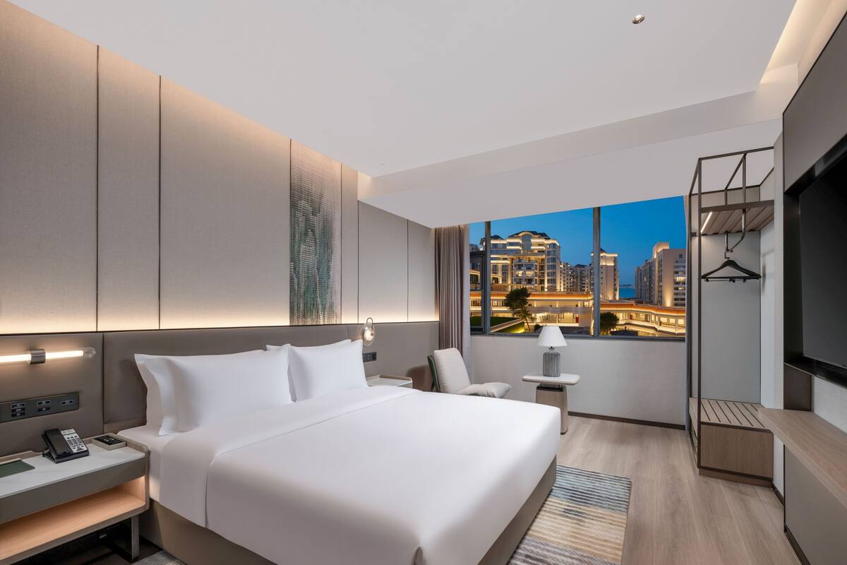 Habitación de hotel moderna con una cama grande, mesitas de noche, un escritorio y vista a un paisaje urbano al anochecer.