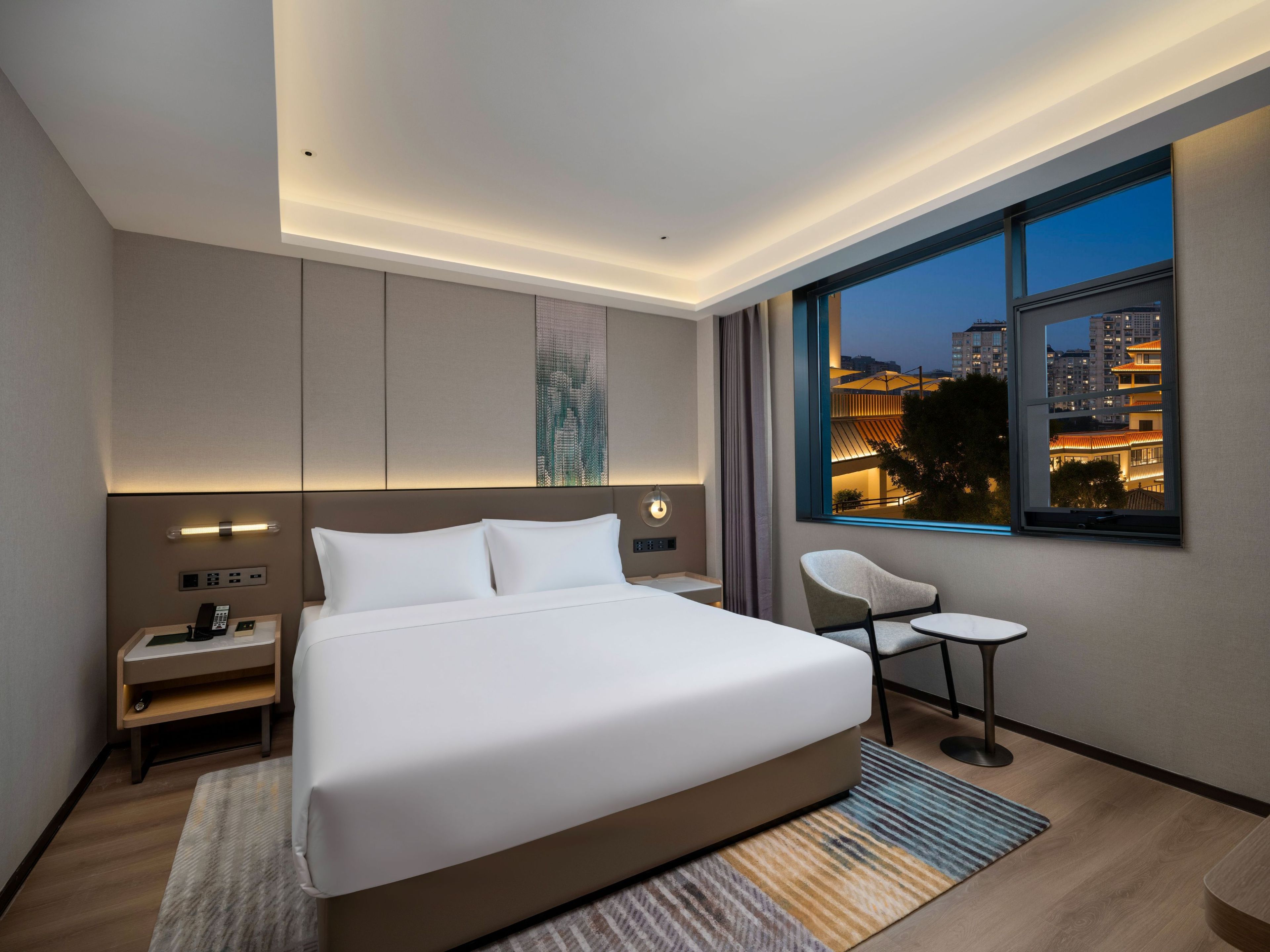 A modern hotel room with a large bed, bedside tables, a chair, and a view of the cityscape at dusk.