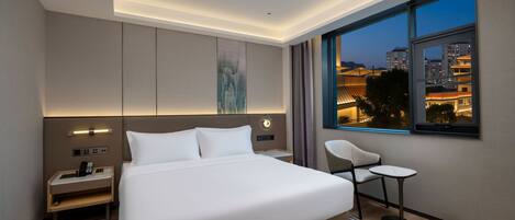 A modern hotel room with a large bed, bedside tables, a chair, and a view of the cityscape at dusk.