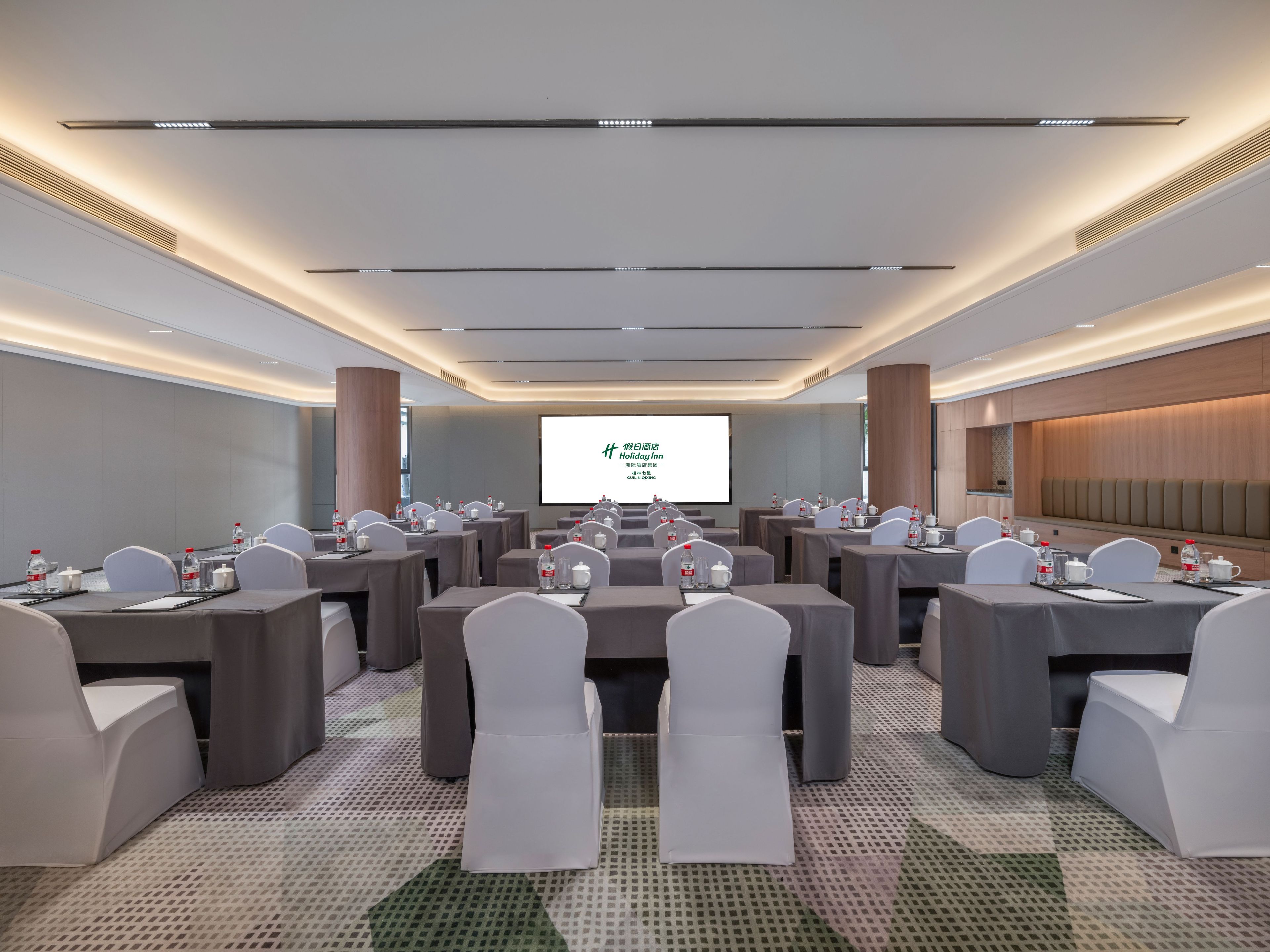 Un salón de conferencias con varias mesas y sillas, una pantalla grande al frente y un diseño de techo moderno.