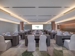 Un salón de conferencias con varias mesas y sillas, una pantalla grande al frente y un diseño de techo moderno.