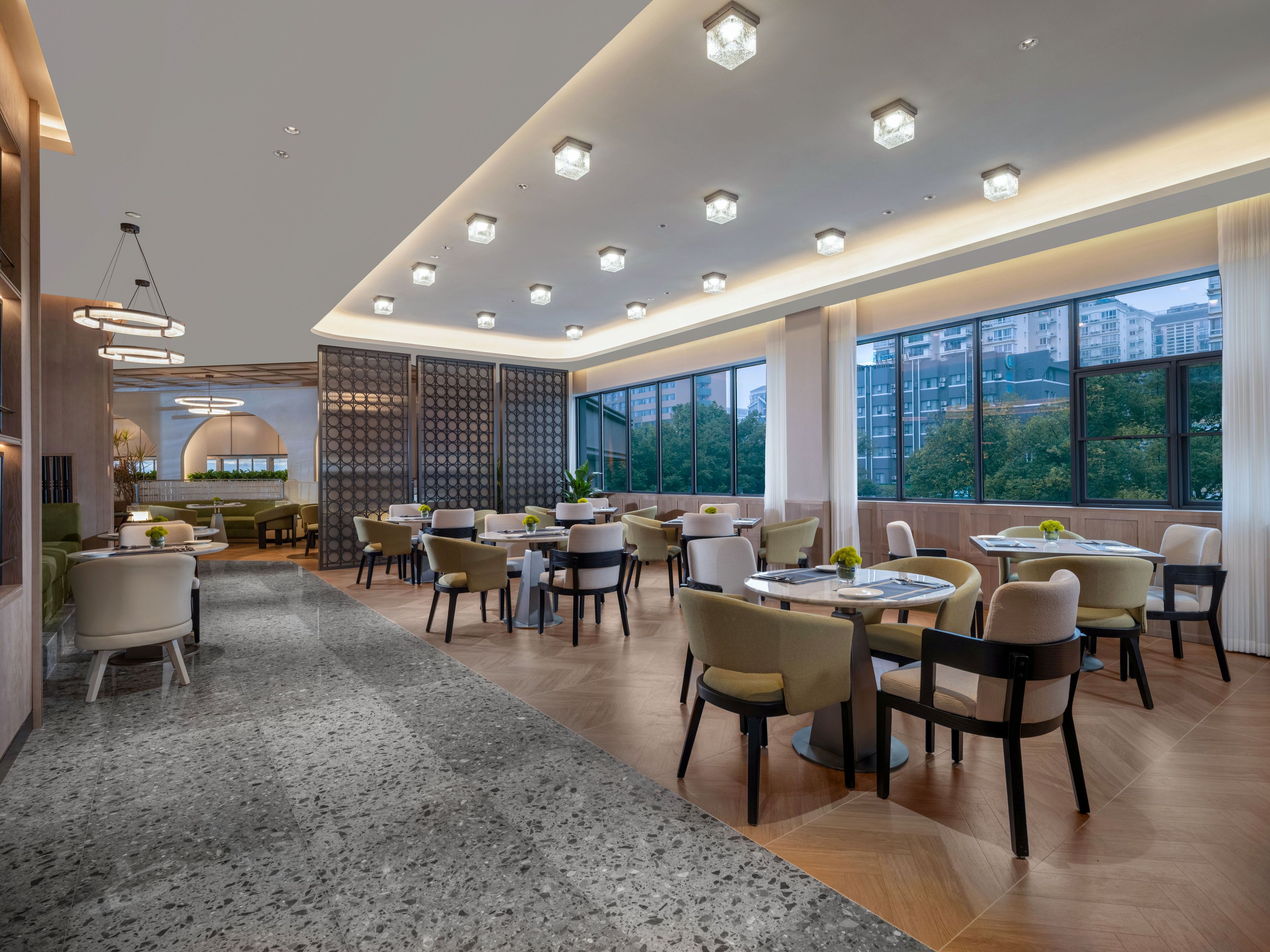 Un comedor amplio con mesas y sillas redondas, ventanas grandes que ofrecen vistas a edificios y árboles, y un diseño de techo moderno con iluminación empotrada.