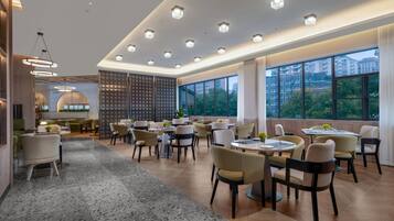 Un comedor amplio con mesas y sillas redondas, ventanas grandes que ofrecen vistas a edificios y árboles, y un diseño de techo moderno con iluminación empotrada.