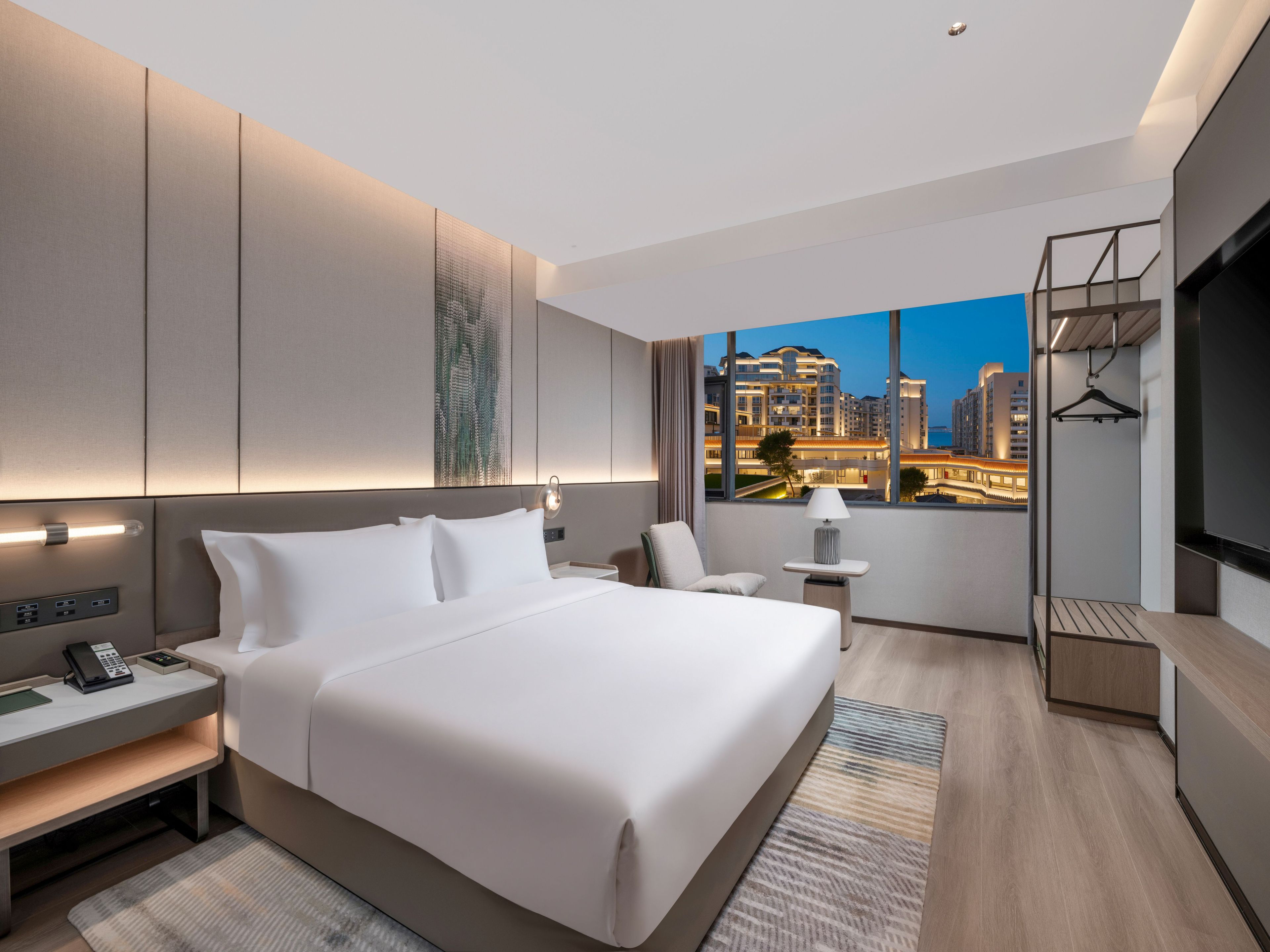 Habitación de hotel moderna con una cama grande, mesitas de noche, un escritorio y vista a un paisaje urbano al anochecer.