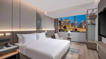 Habitación de hotel moderna con una cama grande, mesitas de noche, un escritorio y vista a un paisaje urbano al anochecer.