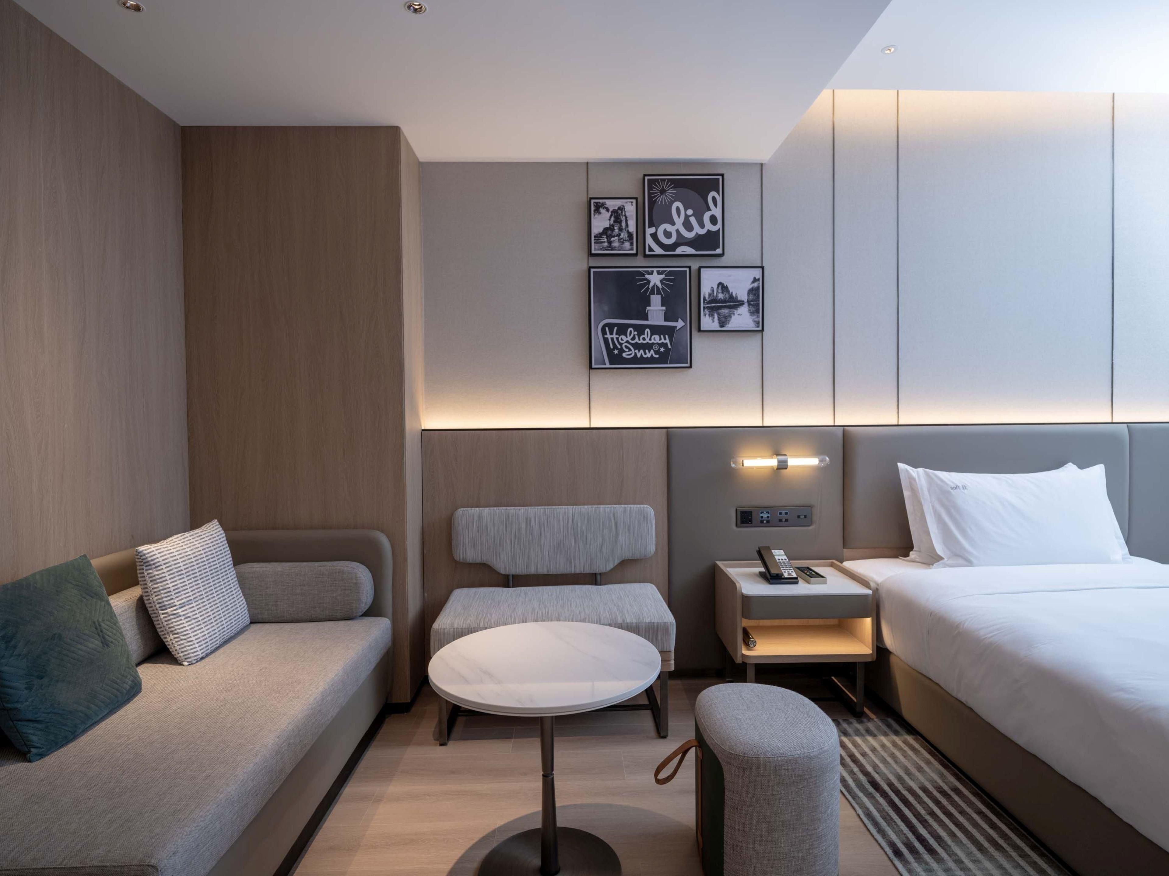 A modern hotel room with a sofa, a round table, a bedside table with a lamp, a bed, and wall-mounted artwork.